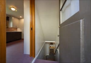 A view from a hallway into a kitchen area with a partially open door revealing a staircase leading down to a basement with shelves and a wine rack.