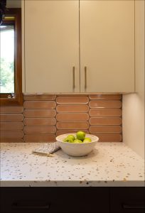 A white bowl with green apples sits on a terrazzo countertop beneath beige cabinets and a window, with a geometric tile backsplash and a folded kitchen towel nearby.