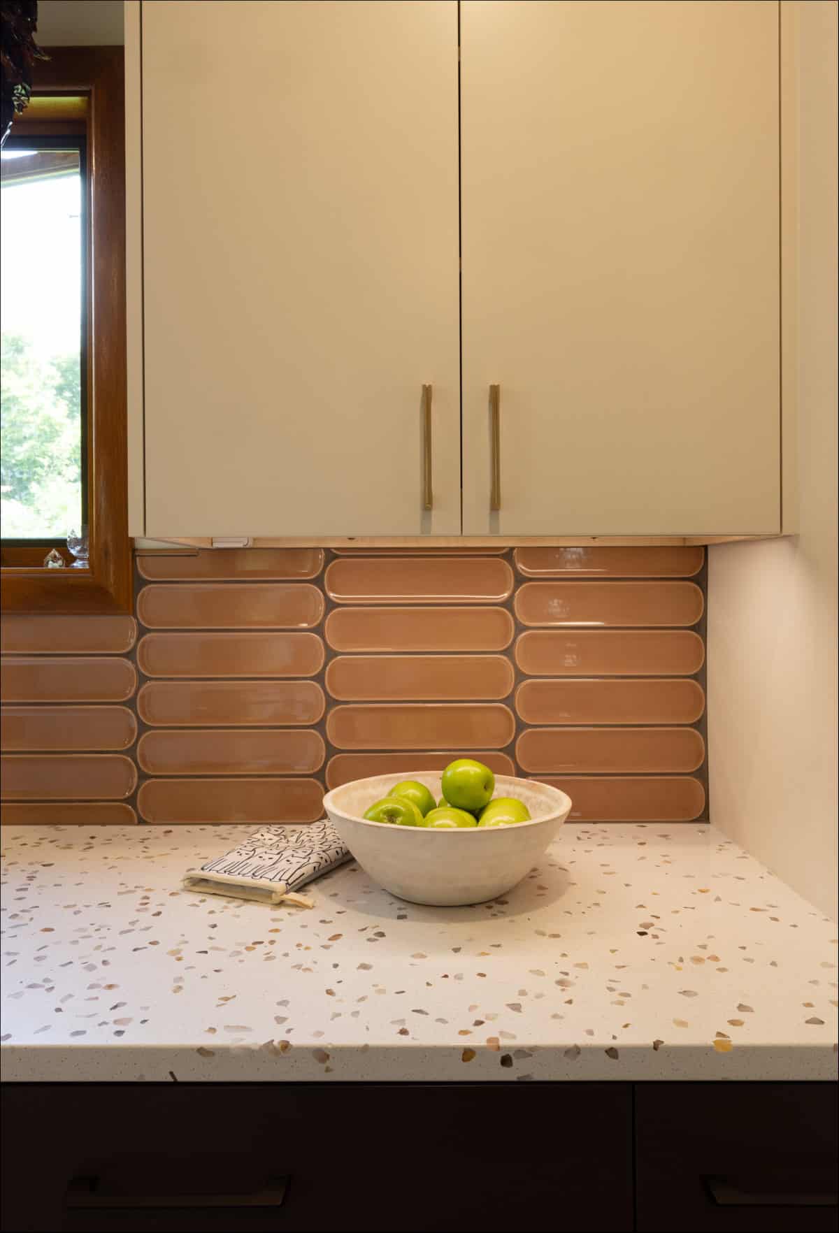 A white bowl with green apples sits on a terrazzo countertop beneath beige cabinets and a window, with a geometric tile backsplash and a folded kitchen towel nearby. A white bowl with green apples sits on a terrazzo countertop beneath beige cabinets and a window, with a geometric tile backsplash and a folded kitchen towel nearby.