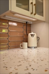 A white electric kettle and a beige mug sit on a speckled countertop beneath a cabinet with gold handles and a brown tiled backsplash.