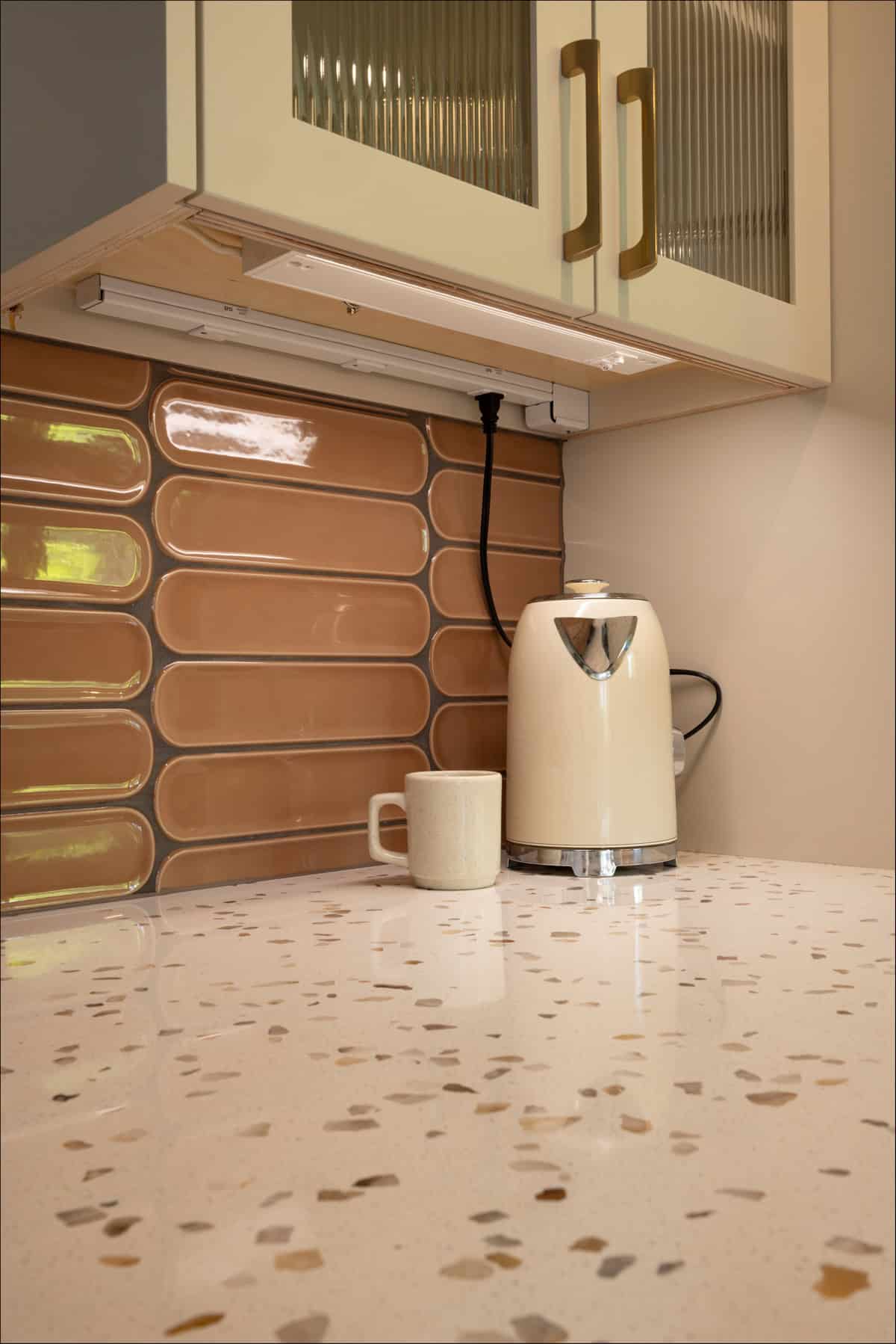 A white electric kettle and a beige mug sit on a speckled countertop beneath a cabinet with gold handles and a brown tiled backsplash. A white electric kettle and a beige mug sit on a speckled countertop beneath a cabinet with gold handles and a brown tiled backsplash.