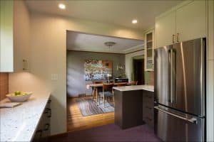 Modern kitchen with stainless steel refrigerator and white cabinets, leading into a dining area with a wooden table and chairs on a striped rug.
