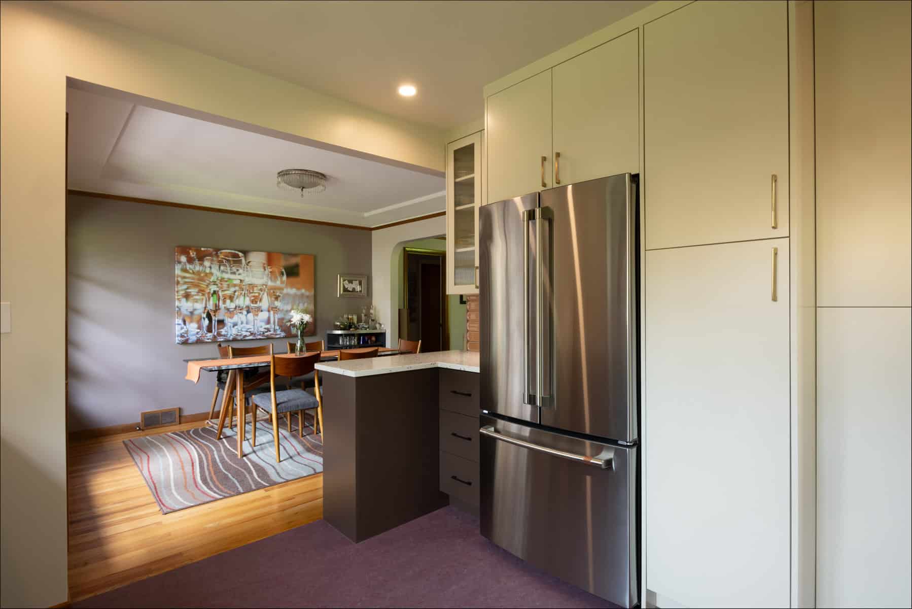 A modern kitchen with stainless steel refrigerator, white cabinets, and a small bar area opens to a dining room with a wooden table and chairs on a striped rug. A modern kitchen with stainless steel refrigerator, white cabinets, and a small bar area opens to a dining room with a wooden table and chairs on a striped rug.