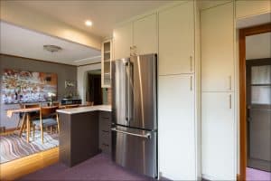 Modern kitchen with stainless steel French door refrigerator, beige cabinets, small island, and view into a dining area with a table and glassware wall art.