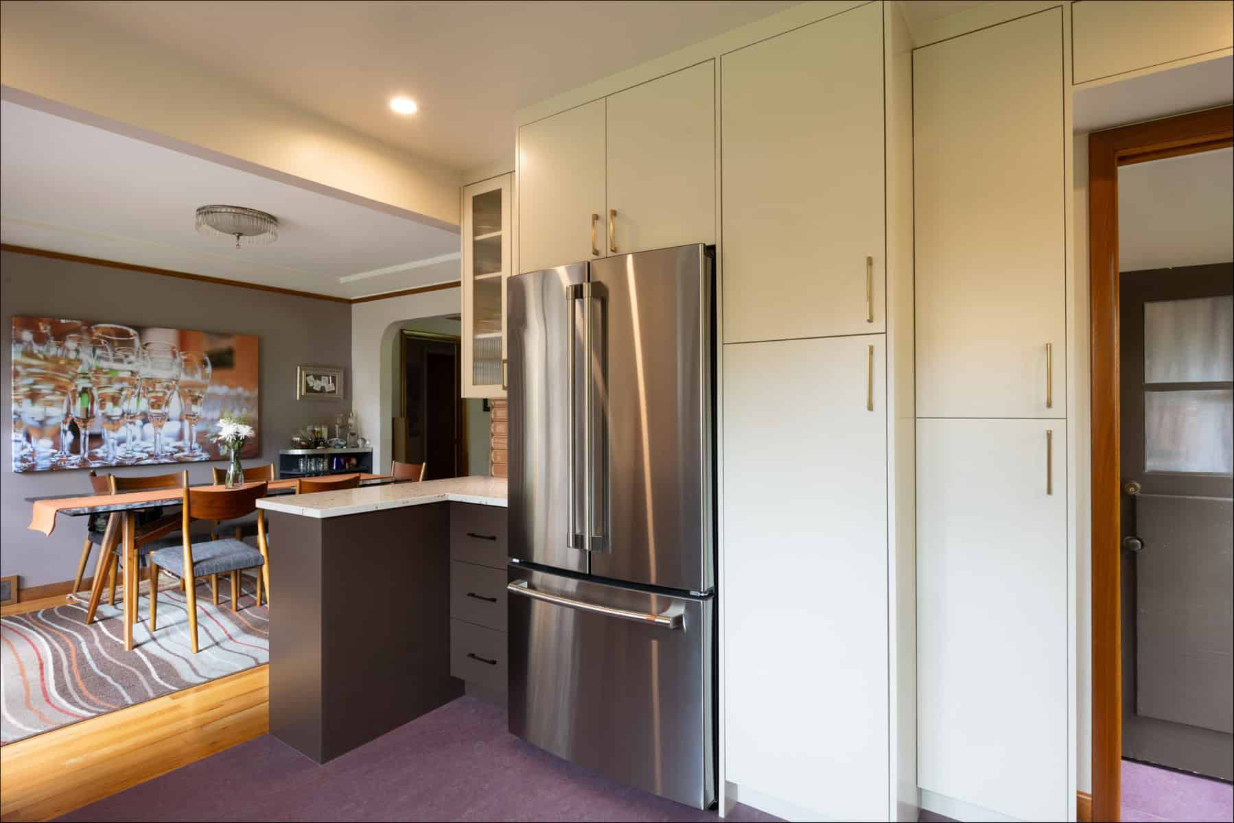 Modern kitchen with stainless steel French door refrigerator, beige cabinets, small island, and view into a dining area with a table and glassware wall art. Modern kitchen with stainless steel French door refrigerator, beige cabinets, small island, and view into a dining area with a table and glassware wall art.