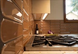A modern kitchen with brown tile backsplash, a gas stove, a bottle of wine, corks, a halved red cabbage, and asparagus on the counter near a window.