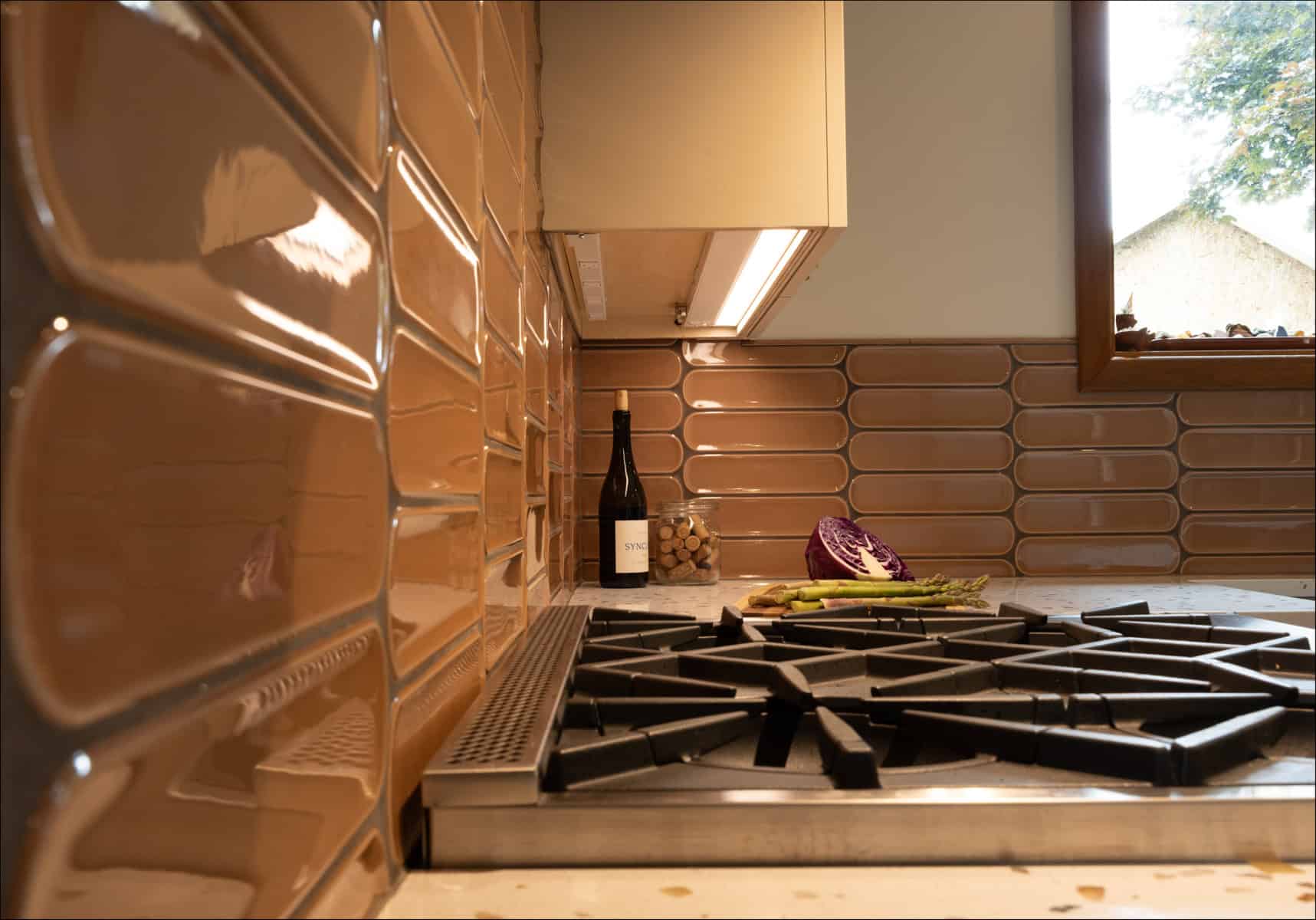 A modern kitchen with brown tile backsplash, a gas stove, a bottle of wine, corks, a halved red cabbage, and asparagus on the counter near a window. A modern kitchen with brown tile backsplash, a gas stove, a bottle of wine, corks, a halved red cabbage, and asparagus on the counter near a window.