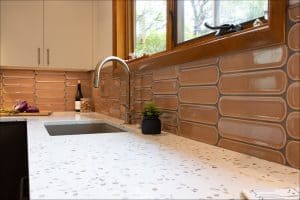 A modern kitchen countertop with a speckled surface, built-in sink, chrome faucet, tan tile backsplash, and a window above the sink.