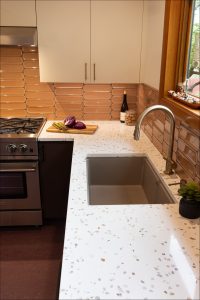 Modern kitchen with a terrazzo countertop, stainless steel sink, and stove. A cutting board with vegetables and a bottle of wine are on the counter; tan tiles line the backsplash.