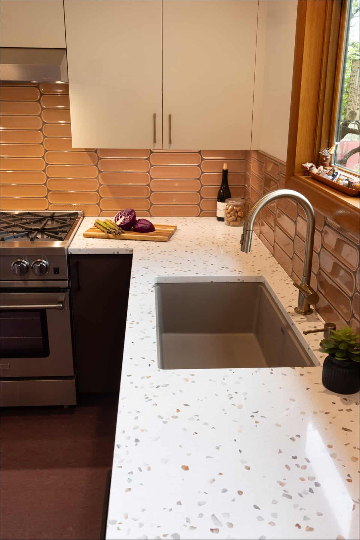 Modern kitchen with a terrazzo countertop, stainless steel sink, and stove. A cutting board with vegetables and a bottle of wine are on the counter; tan tiles line the backsplash. Modern kitchen with a terrazzo countertop, stainless steel sink, and stove. A cutting board with vegetables and a bottle of wine are on the counter; tan tiles line the backsplash.
