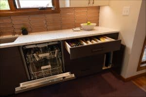 A modern kitchen with an open dishwasher and a drawer containing organized utensils under a white countertop with a bowl of apples.
