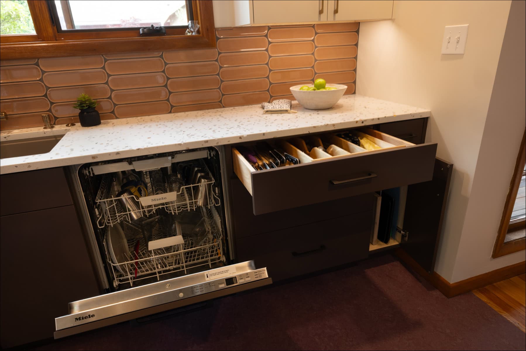 A modern kitchen with an open dishwasher and a drawer containing organized utensils under a white countertop with a bowl of apples. A modern kitchen with an open dishwasher and a drawer containing organized utensils under a white countertop with a bowl of apples.