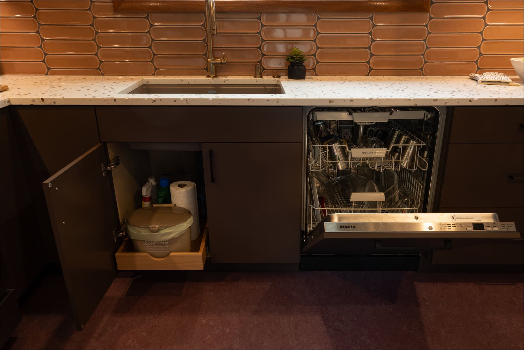 A modern kitchen with a sink, an open dishwasher, and a cabinet door open to reveal cleaning supplies below the counter. A modern kitchen with a sink, an open dishwasher, and a cabinet door open to reveal cleaning supplies below the counter.