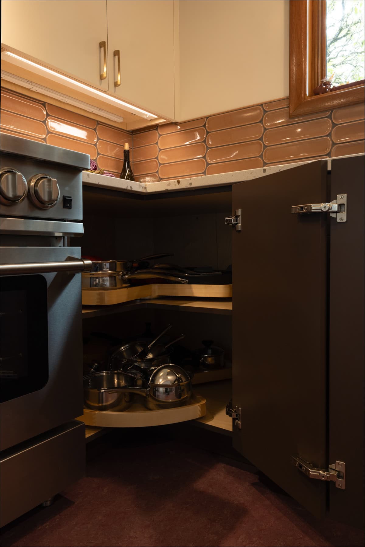Open corner kitchen cabinet with rotating shelves holding various pots and pans, next to a stainless steel oven and under a counter with tile backsplash. Open corner kitchen cabinet with rotating shelves holding various pots and pans, next to a stainless steel oven and under a counter with tile backsplash.