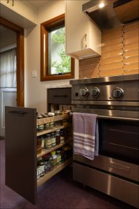 A kitchen with a pull-out spice rack next to a stainless steel oven and stove, with a purple towel hanging on the oven handle and a window above the countertop.