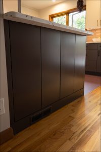 Dark brown kitchen island with a light countertop, air vent at the bottom, and wood flooring in a well-lit room with a window in the background.