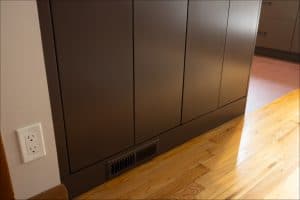 A close-up view of a brown cabinet base with a floor vent and an electrical outlet on the adjacent wall, next to a hardwood floor.