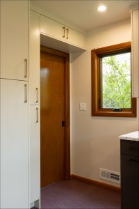 A small room corner with a wooden door, white cabinets, a window with a view of greenery, and recessed lighting.