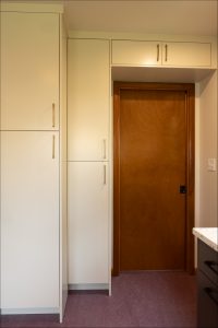 A wooden door is set into a wall next to tall, light-colored cabinets with metal handles in a kitchen or utility room.