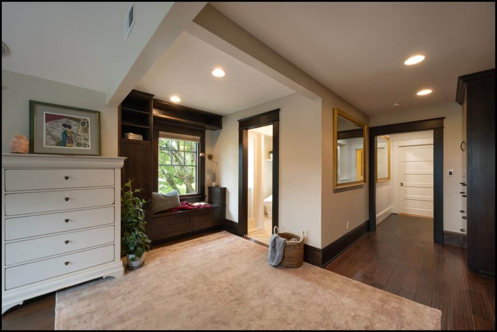 A bedroom with dark wood accents, a window seat, a white dresser, a large mirror, a beige rug, and hardwood floors, with a hallway leading to white doors in the background.