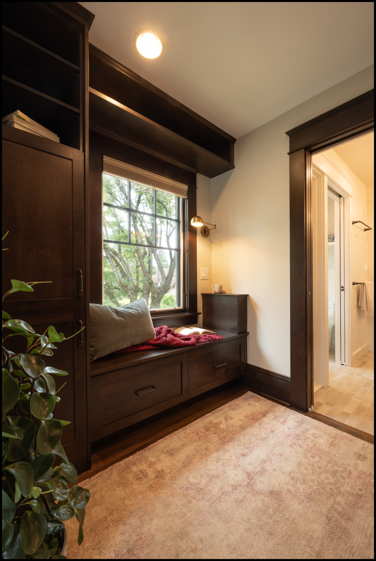 A cozy reading nook with built-in dark wood bench and shelves sits by a window; a doorway leads to a brightly lit bathroom.