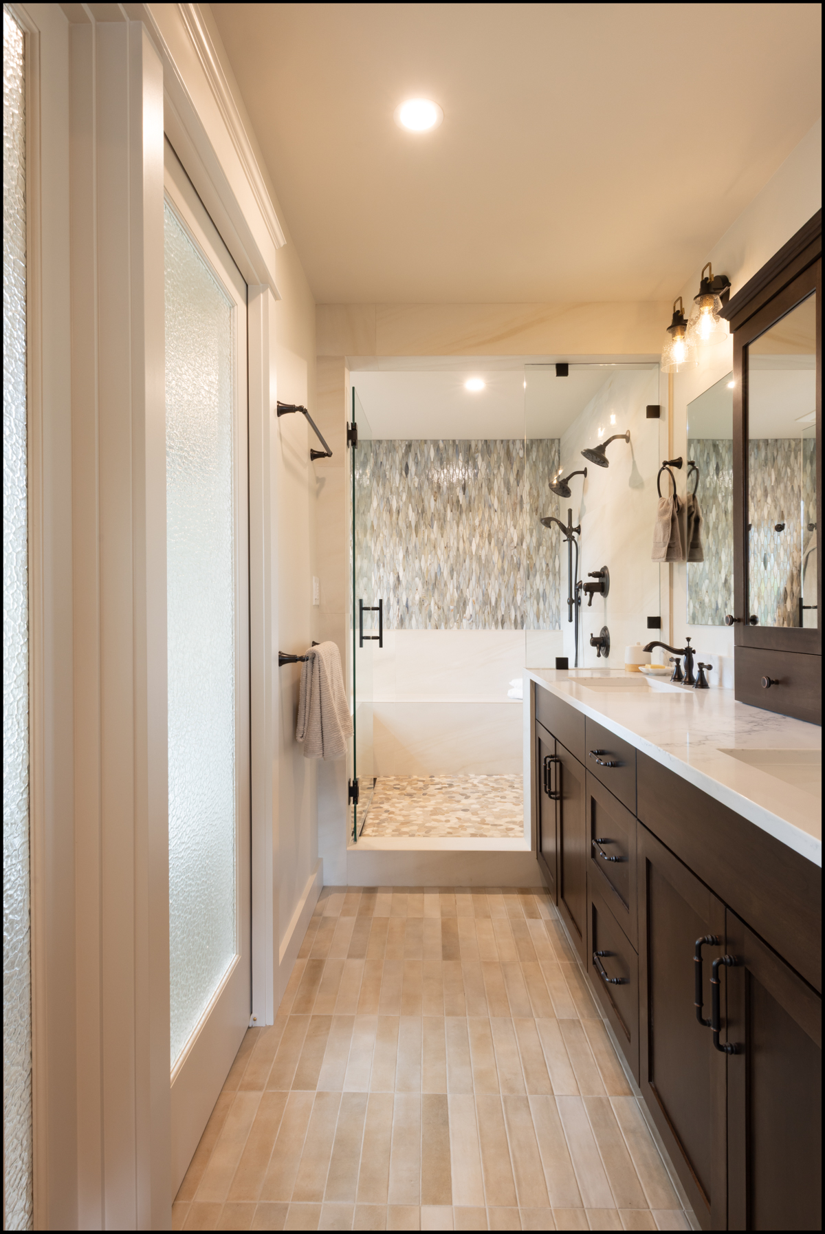 Modern bathroom featuring a glass shower enclosure, a long vanity with dual sinks and dark cabinets, light tile flooring, and patterned shower wall tiles.