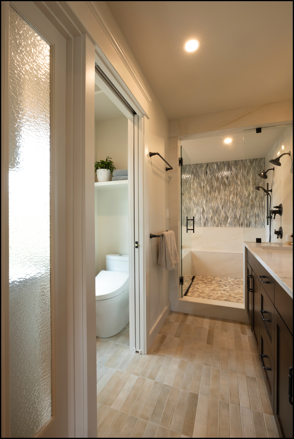 Modern bathroom with a glass shower, built-in shelving above the toilet, and a double vanity with dark cabinets and light countertops.