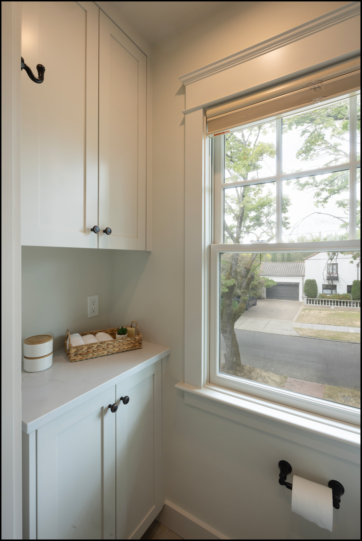 A small bathroom corner with white cabinets, a countertop with a wicker basket and container, a window with a view outside, and a wall-mounted toilet paper holder.