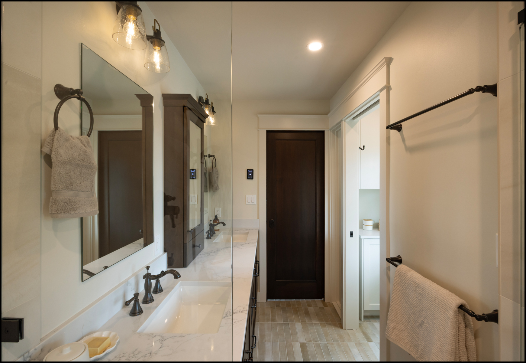 Modern bathroom with dual sinks, marble countertop, wall-mounted lights, mirrors, towel racks, and a dark wooden door leading to an adjacent room with shelving.