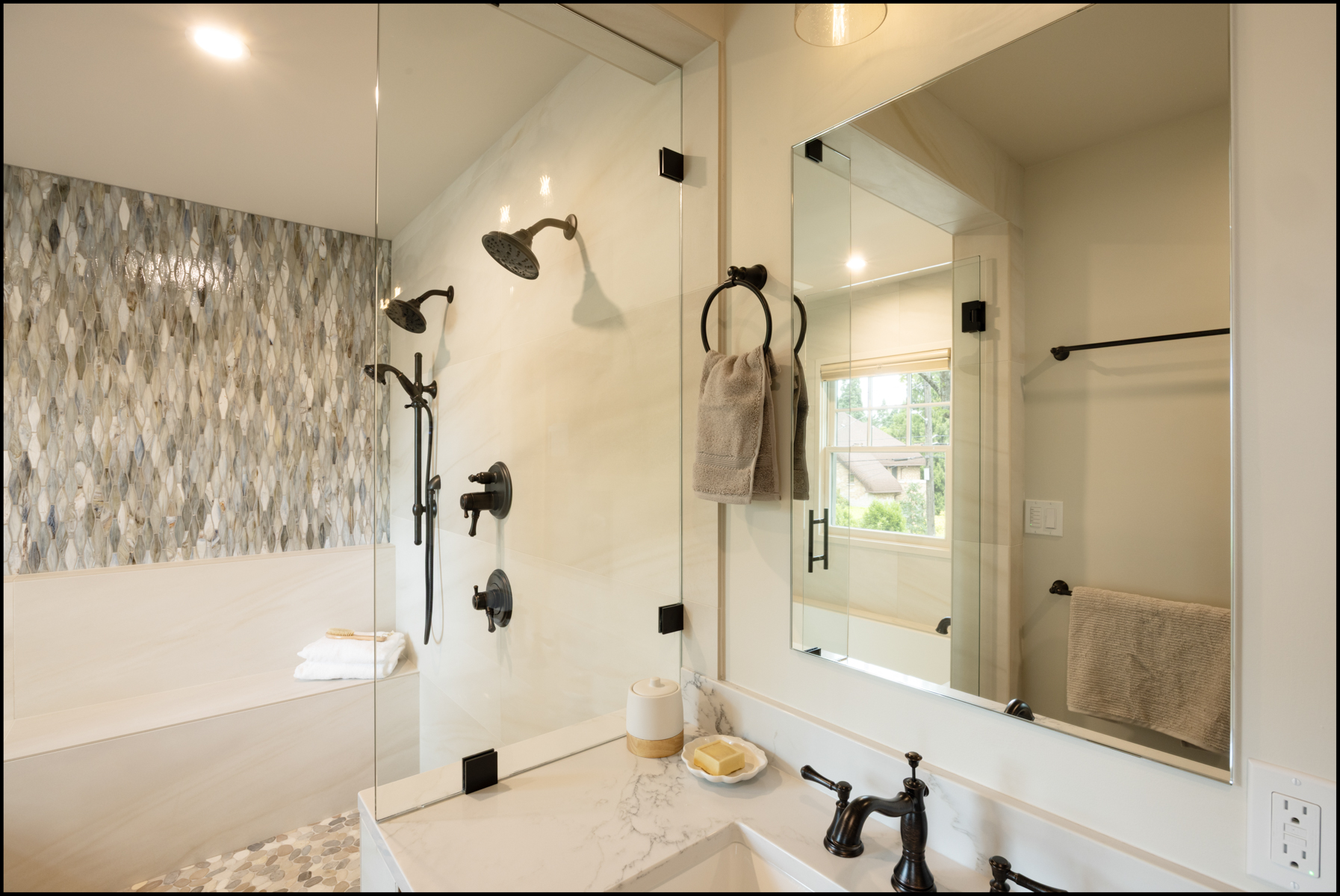 Modern bathroom with glass shower, marble countertop, black fixtures, wall-mounted towel ring, and textured accent wall near a bench with folded towels.