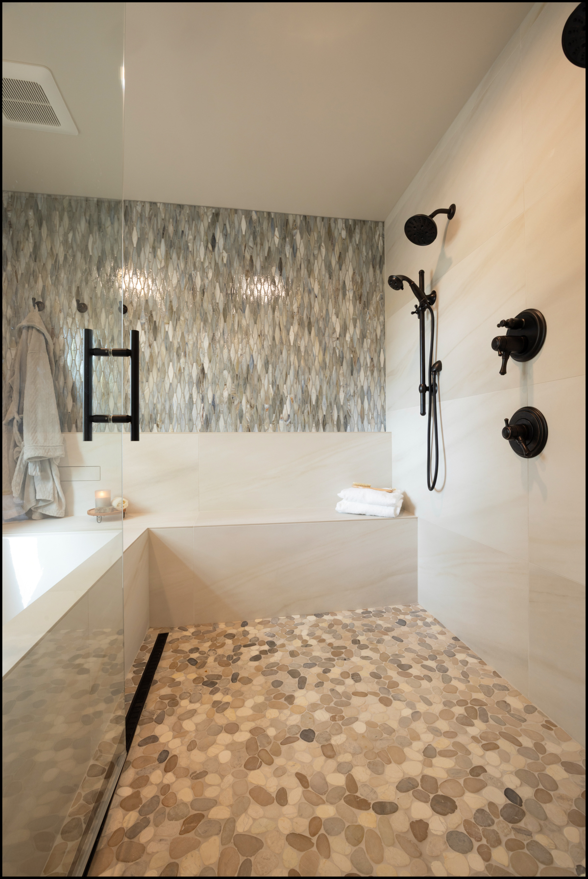 Modern bathroom with a pebble-tiled shower floor, black fixtures, a built-in bathtub, and a patterned accent wall with hanging towels.