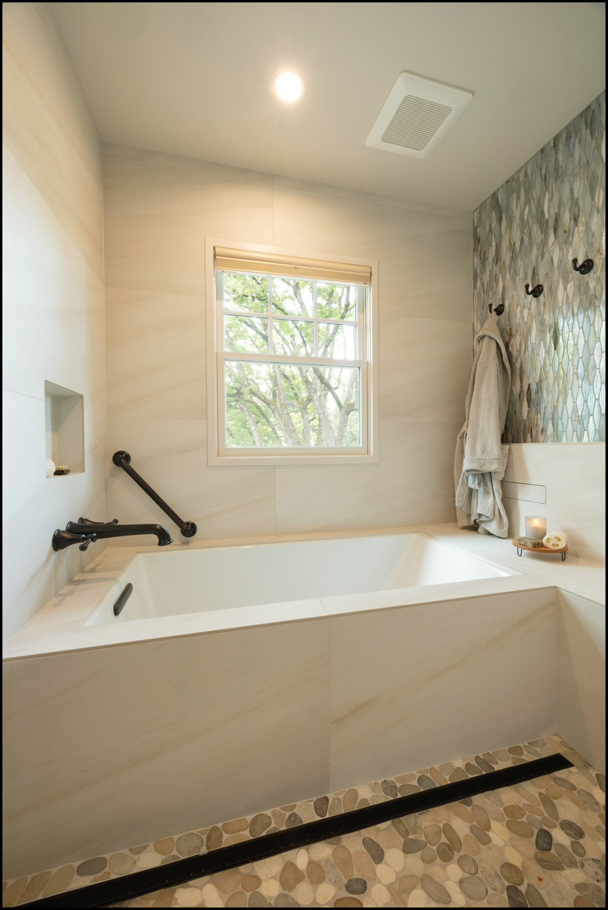 Modern bathroom with a rectangular bathtub, pebble stone floor, wall-mounted grab bars, a window, towel hooks, and small shelf with bath items.