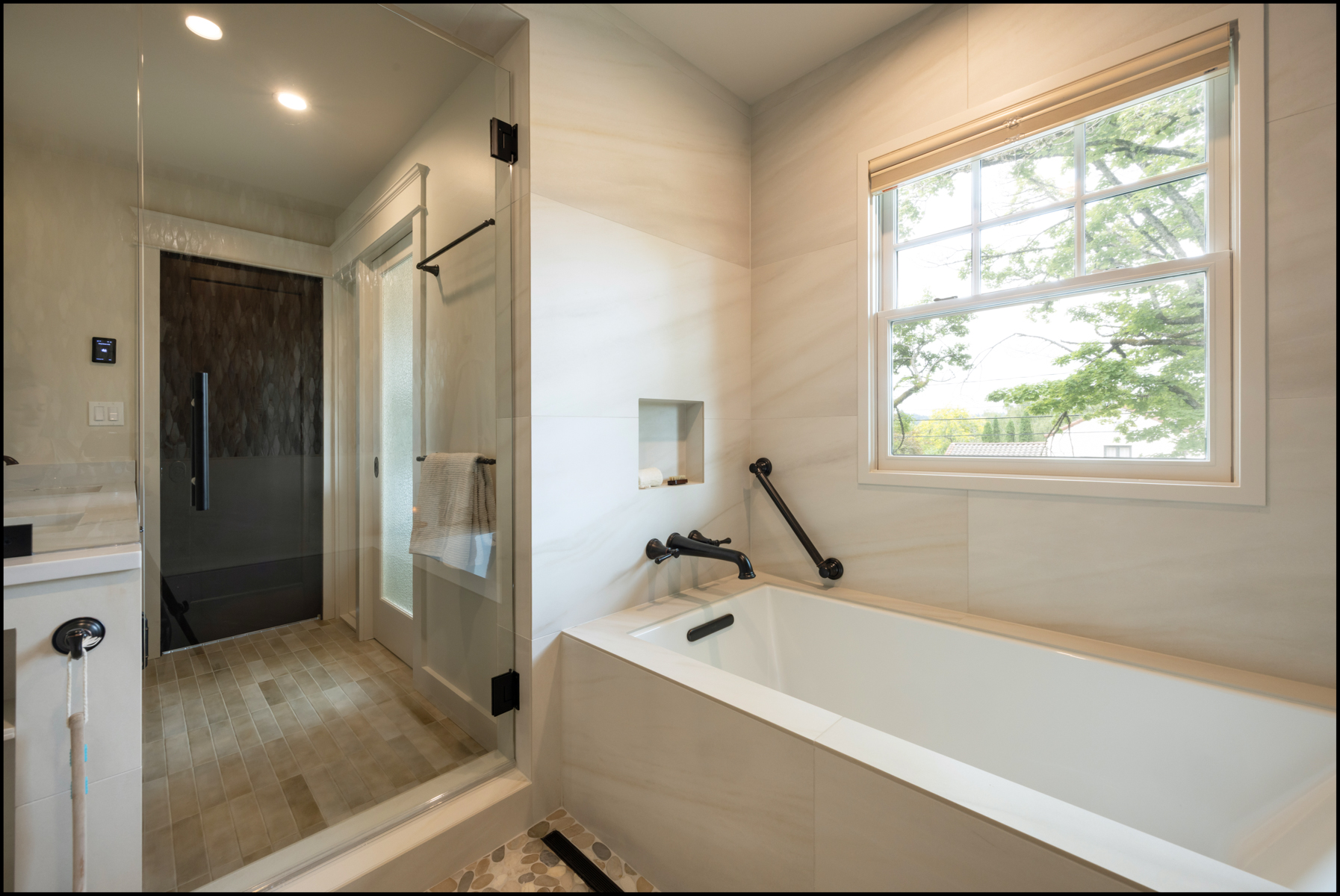 Modern bathroom with a large bathtub, a walk-in shower with glass door, tiled floor, window with outdoor view, and neutral-colored walls.