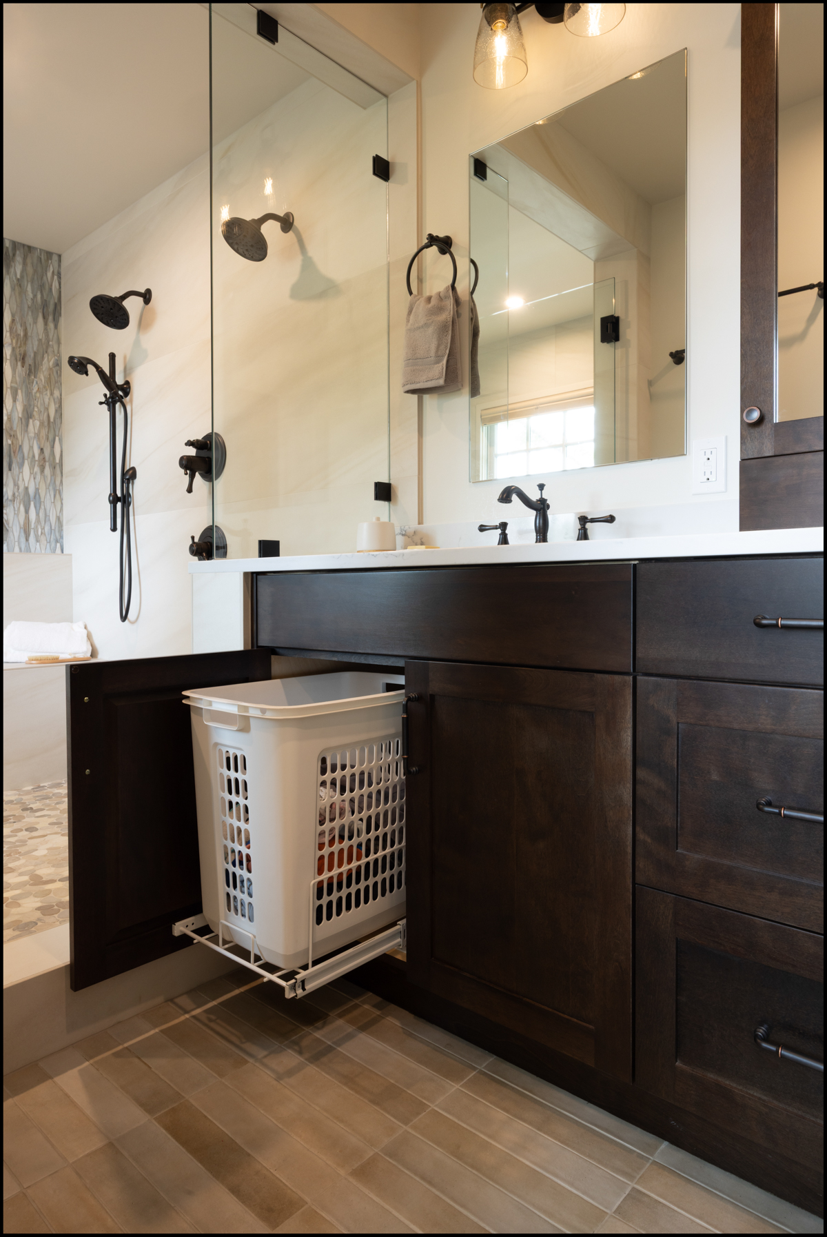Modern bathroom with dark wood cabinets, dual sink, black fixtures, glass shower, and a pull-out laundry hamper partially open beneath the counter.