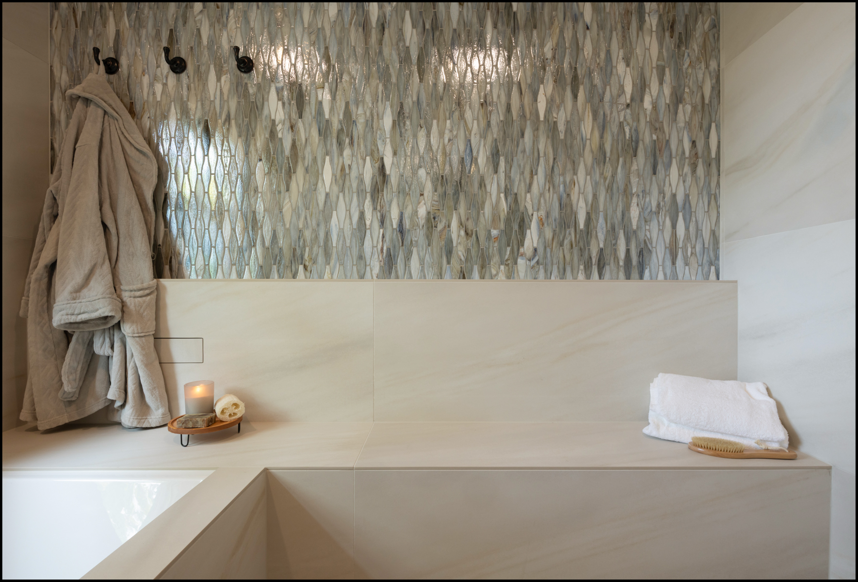 A bathroom with a light stone bench, a folded towel, a wooden brush, a lit candle, a bathrobe on hooks, and a decorative patterned tile wall.