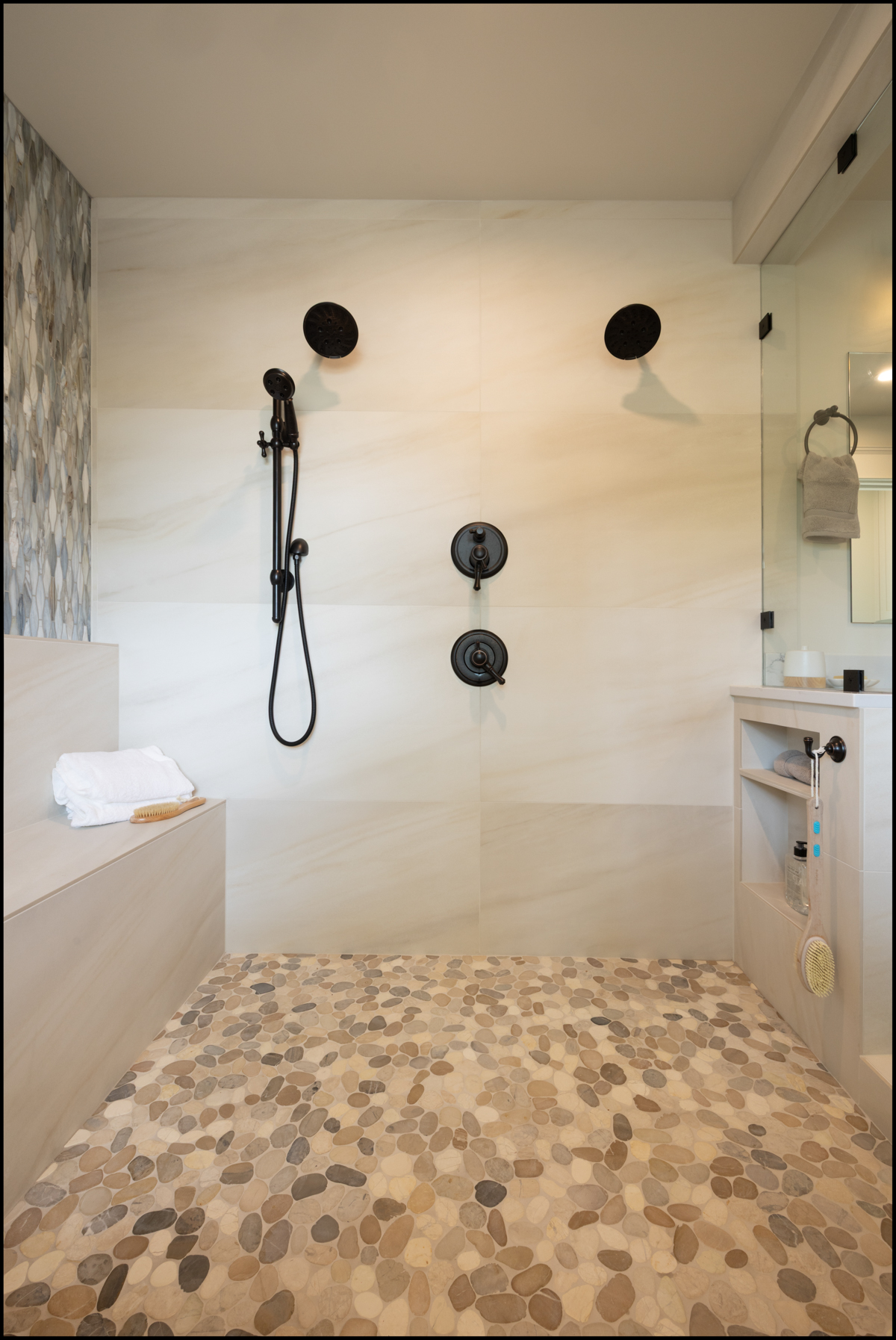 Modern walk-in shower with dual black shower heads, built-in bench, pebble-patterned floor, niche shelves, and neutral-toned wall tiles.