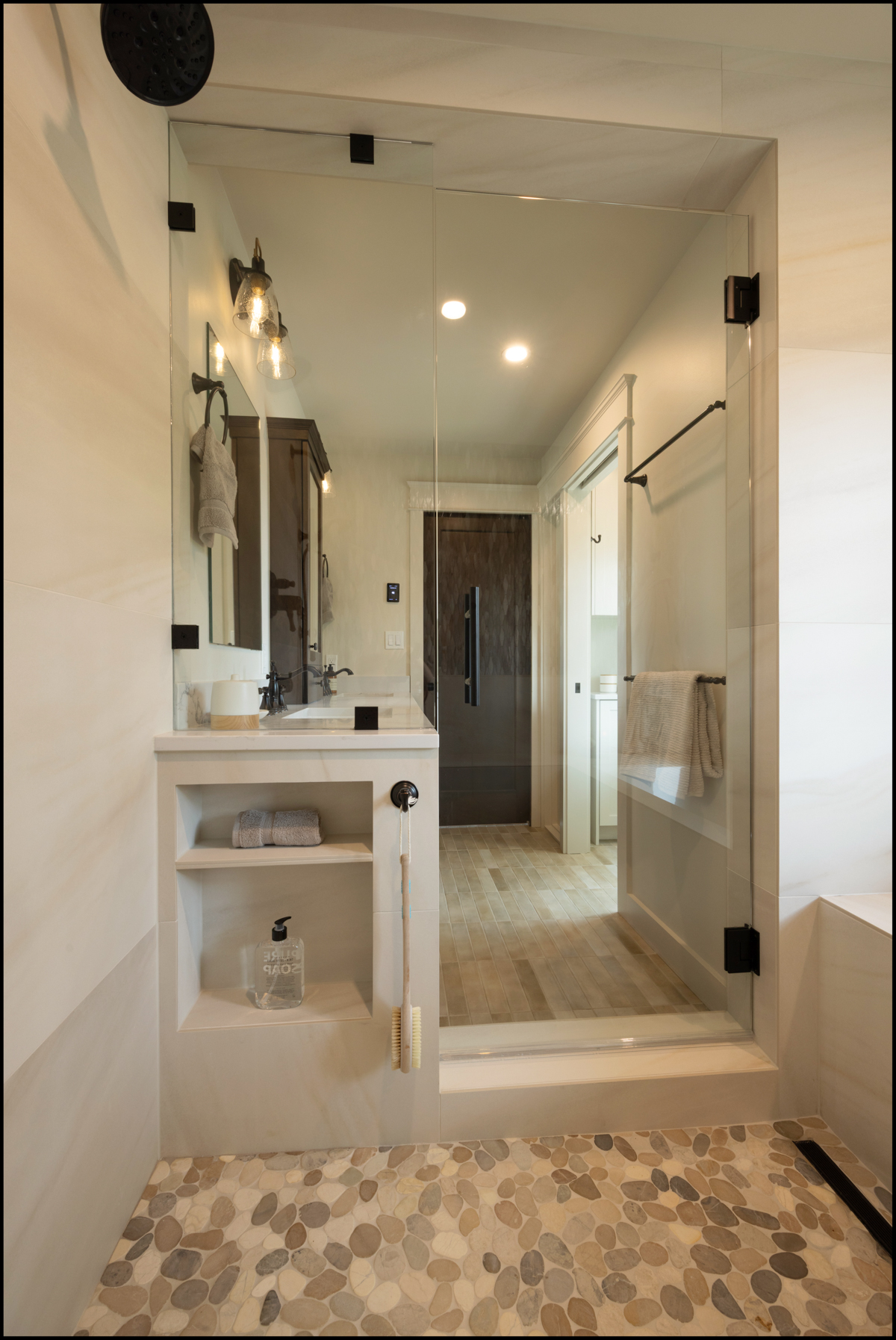 Modern bathroom with a glass-enclosed shower, pebble stone floor, built-in shelves, towel bar, and view into a vanity area with lights and mirrors.