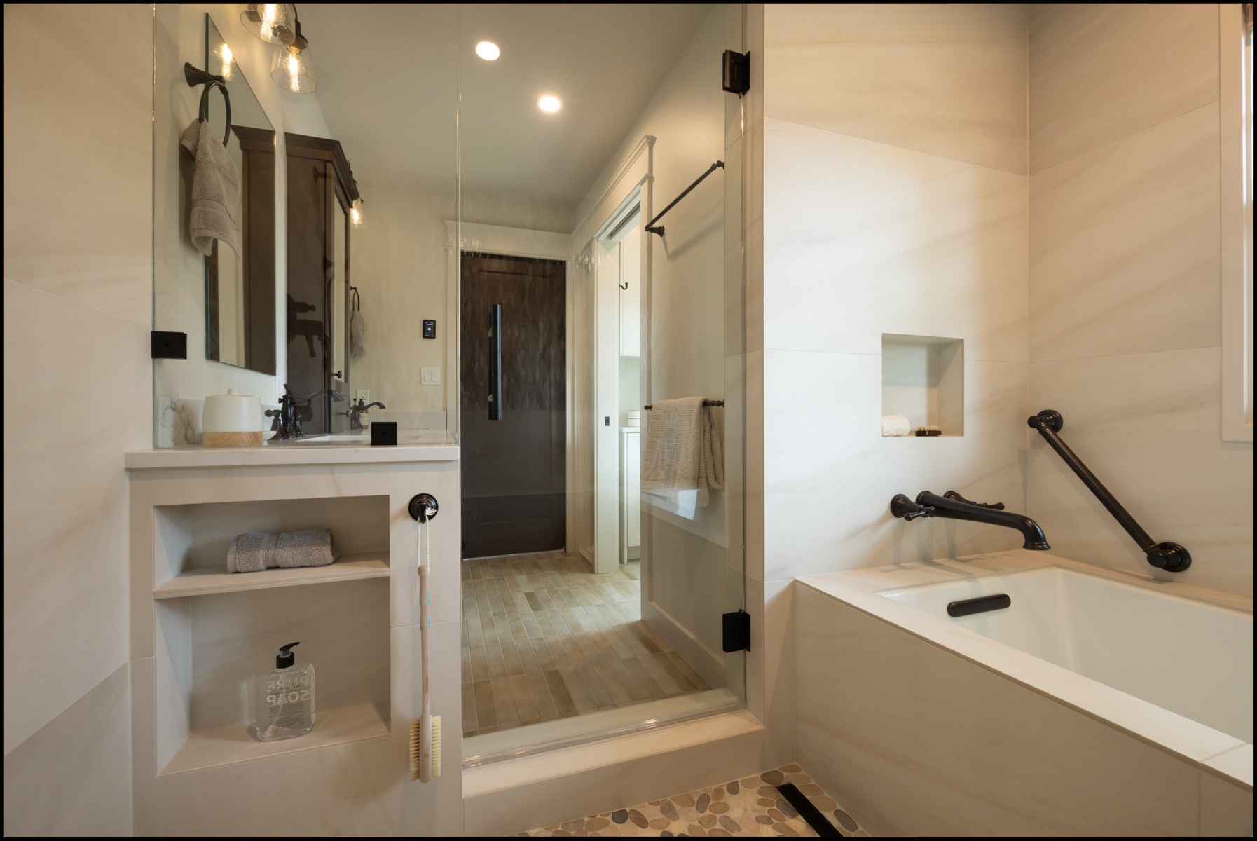 Modern bathroom with a glass shower, built-in shelving, a bathtub with grab bars, neutral tiles, and a towel hanging on a wall-mounted bar.