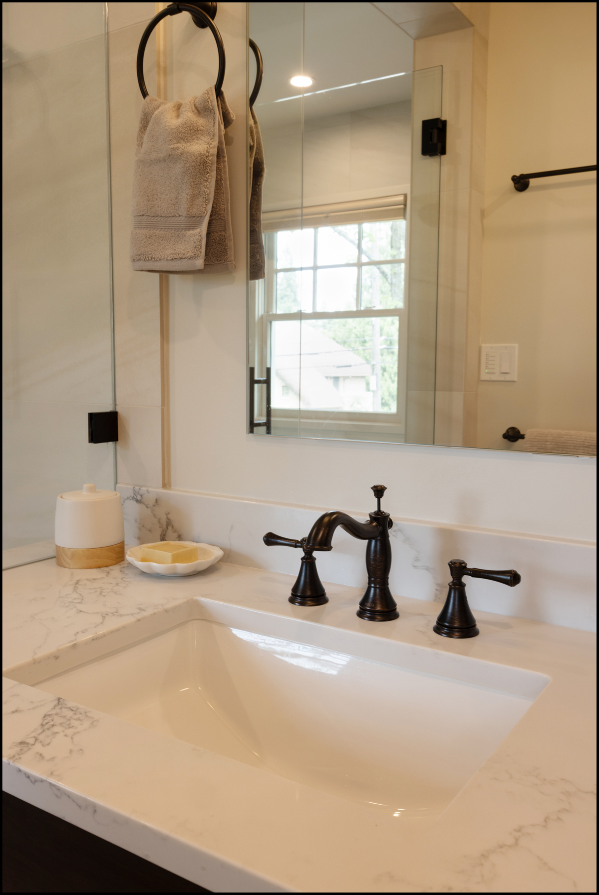A modern bathroom sink with a marble countertop, black faucet, soap dish, tissue holder, and two beige towels hanging on a ring next to a mirror.