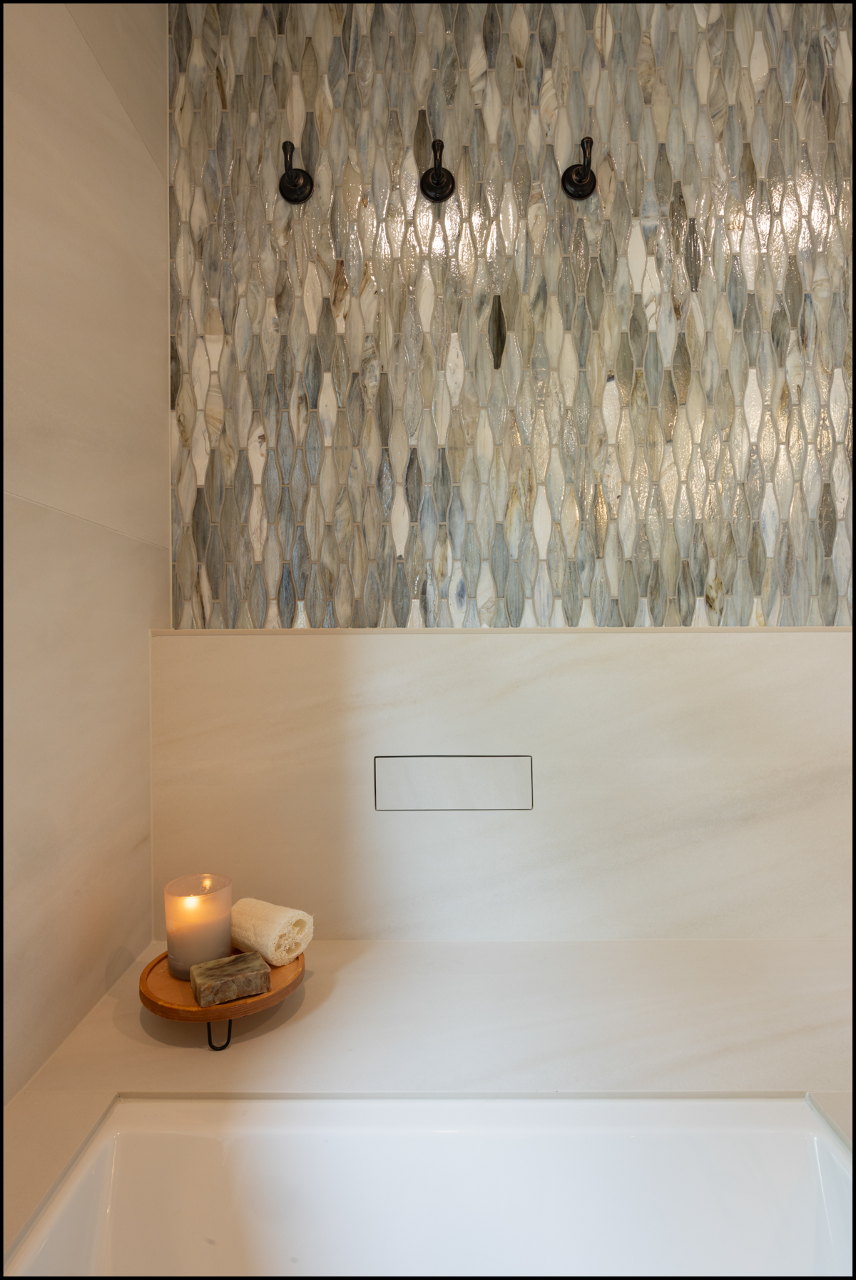 A bathroom with a patterned tile wall, three black hooks, and a countertop holding a lit candle, rolled towel, and soap on a wooden tray.