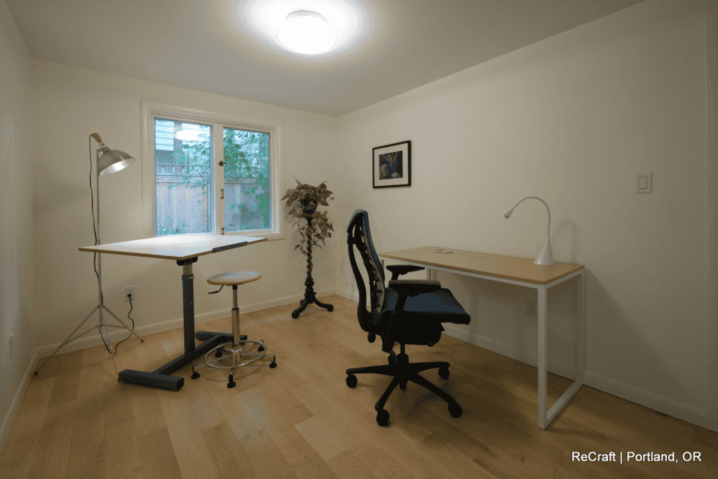 A minimalist home office, perfect for a family basement remodel, with a desk, office chair, lamp, adjustable stool, potted plant, and a window providing natural light.