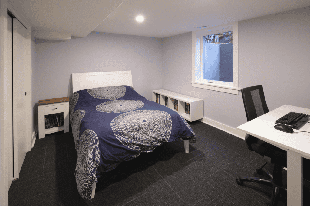 A small bedroom with a bed, nightstand, bookshelf, and desk with a computer chair—perfect for a basement renovation—featuring a window and neutral-colored walls and flooring.