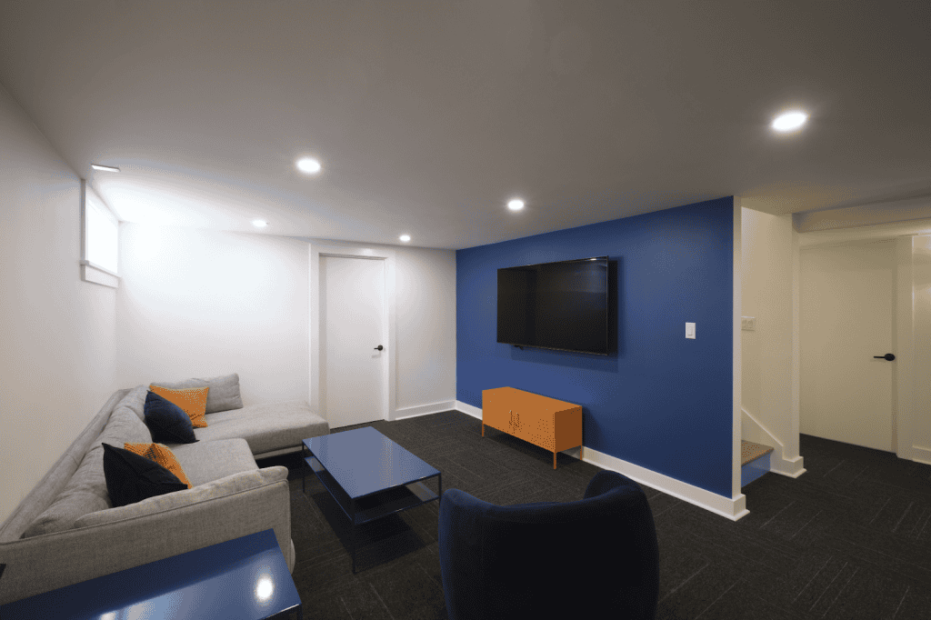 Modern basement living room with a gray sectional sofa, blue accent wall, mounted TV, blue and orange furniture, and recessed ceiling lights—perfect inspiration for your next basement remodel.