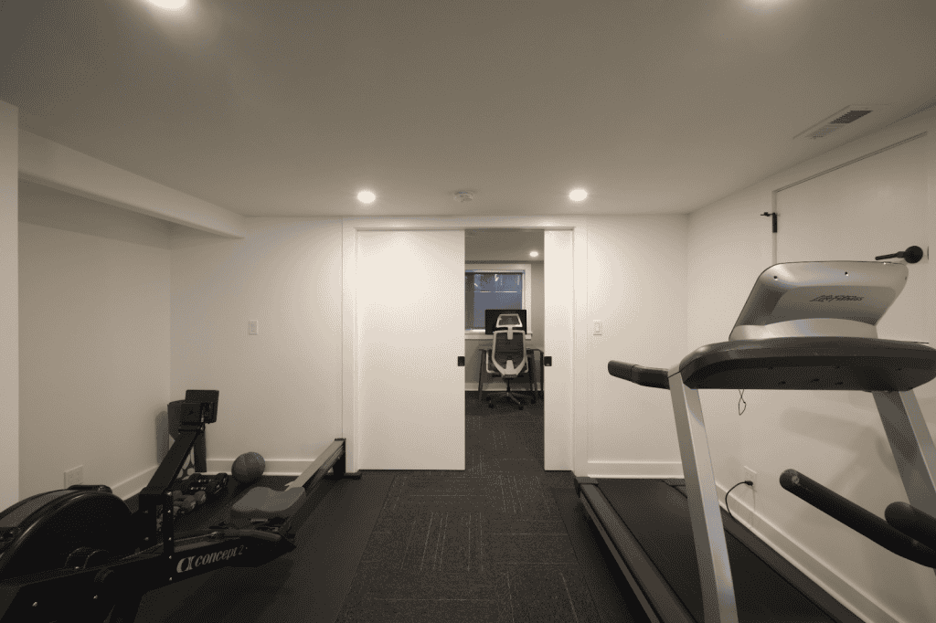 A family-friendly remodel features a home gym room with a treadmill, rowing machine, exercise ball, and a view into an adjacent office space with a desk and chair—perfect for any basement renovation.