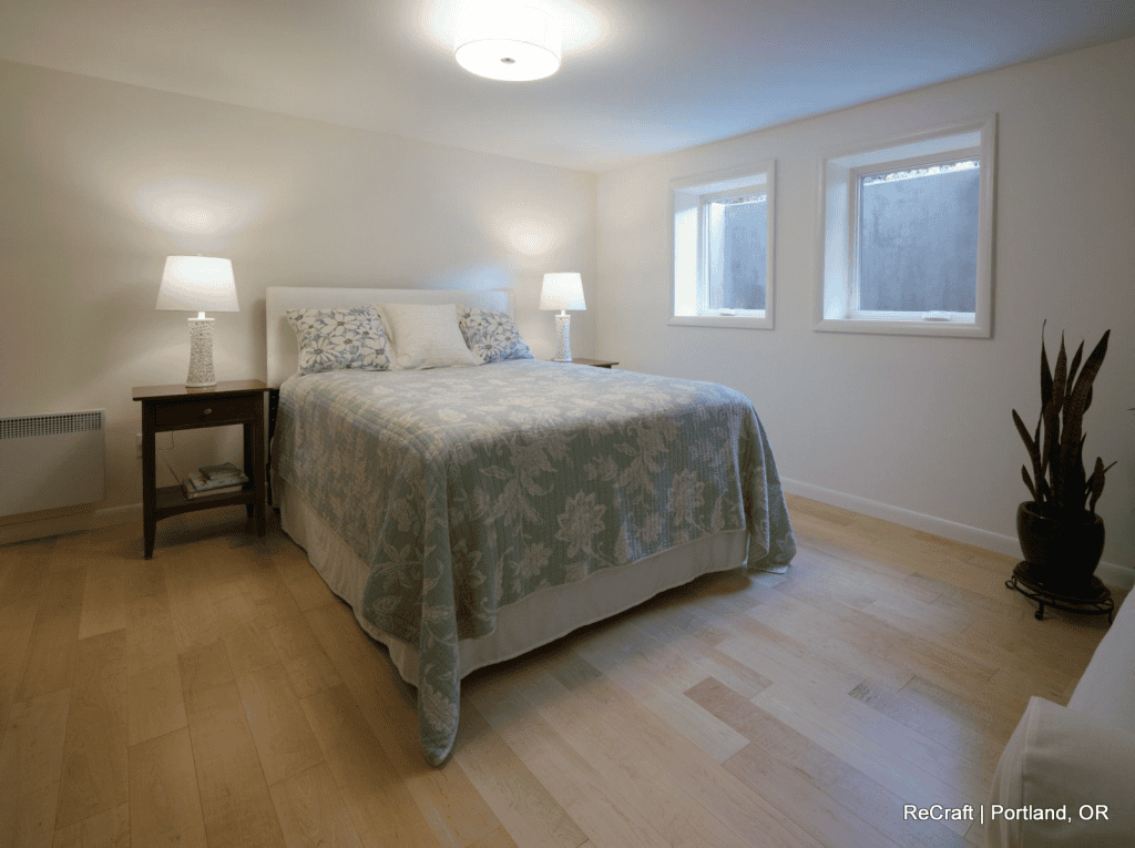A simple bedroom from a recent basement remodel, featuring light wood floors, a bed with a floral comforter, two bedside tables with lamps, two windows, and a potted plant in the corner.
