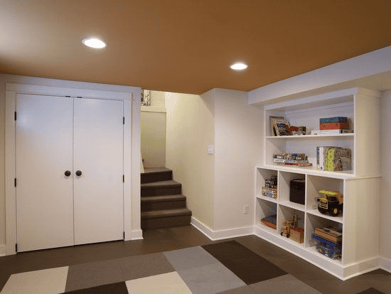 Family-friendly basement room with a tan ceiling, built-in shelves holding toys and games, a double-door closet, carpet tiles on the floor, and stairs leading up—perfect for your next basement remodel.
