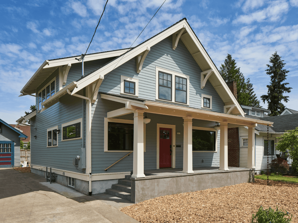 A two-story blue craftsman-style house with a covered front porch, white trim, and a gable roof, set against a partly cloudy sky.
