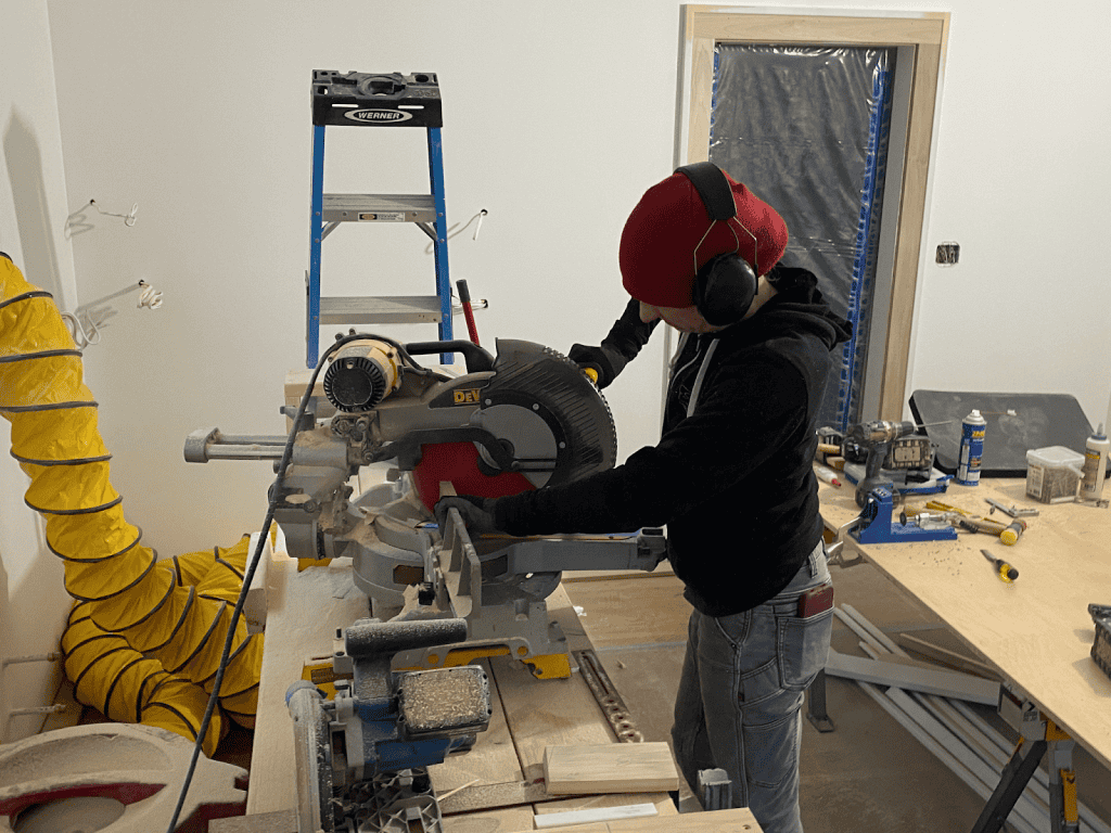 Person wearing a red beanie and ear protection operates a miter saw on a workbench in a construction or renovation setting. Tools and materials are scattered around.