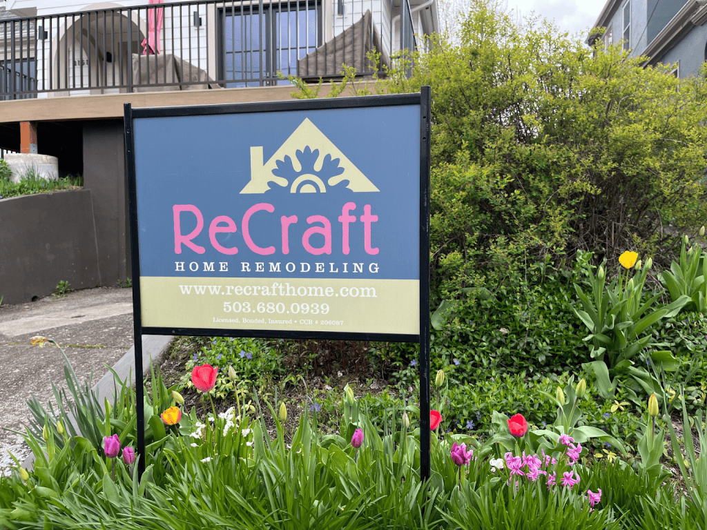 A ReCraft Home Remodeling sign stands in a garden with tulips, displaying the company logo, website, and phone number in front of a residential building.