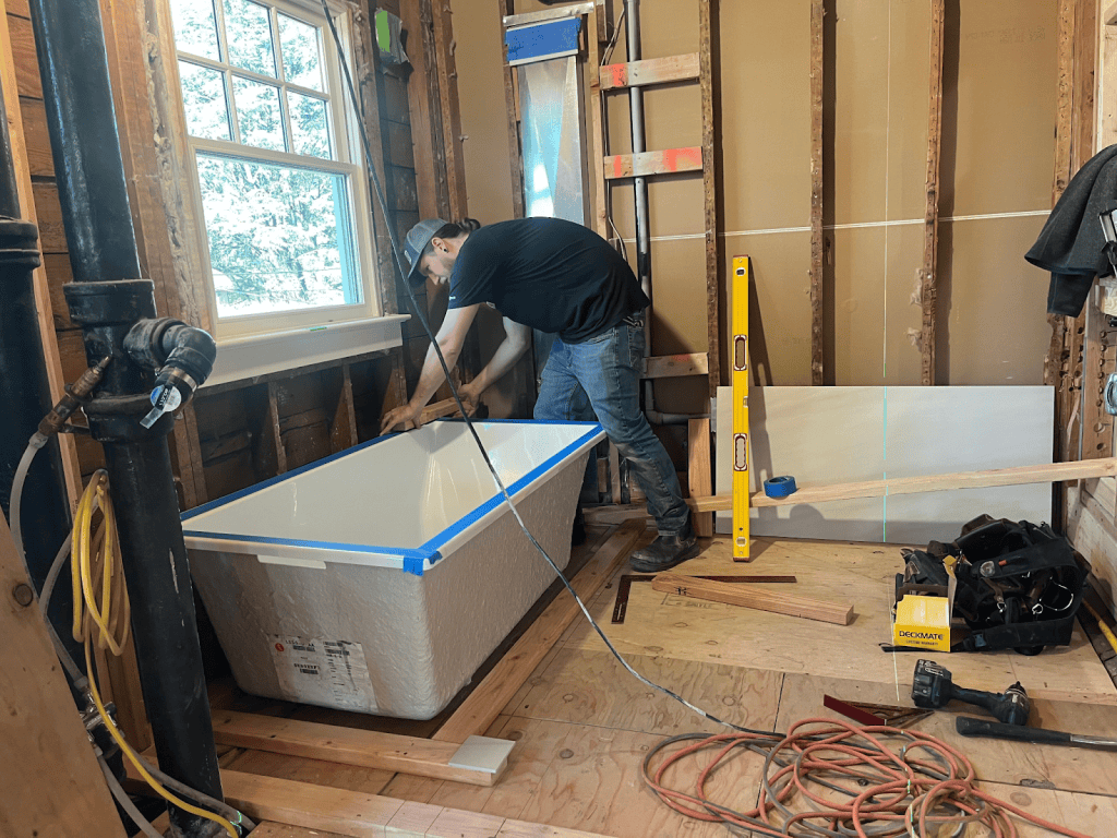 A person installs a bathtub in a partially renovated bathroom with exposed wall studs and various tools on the floor.